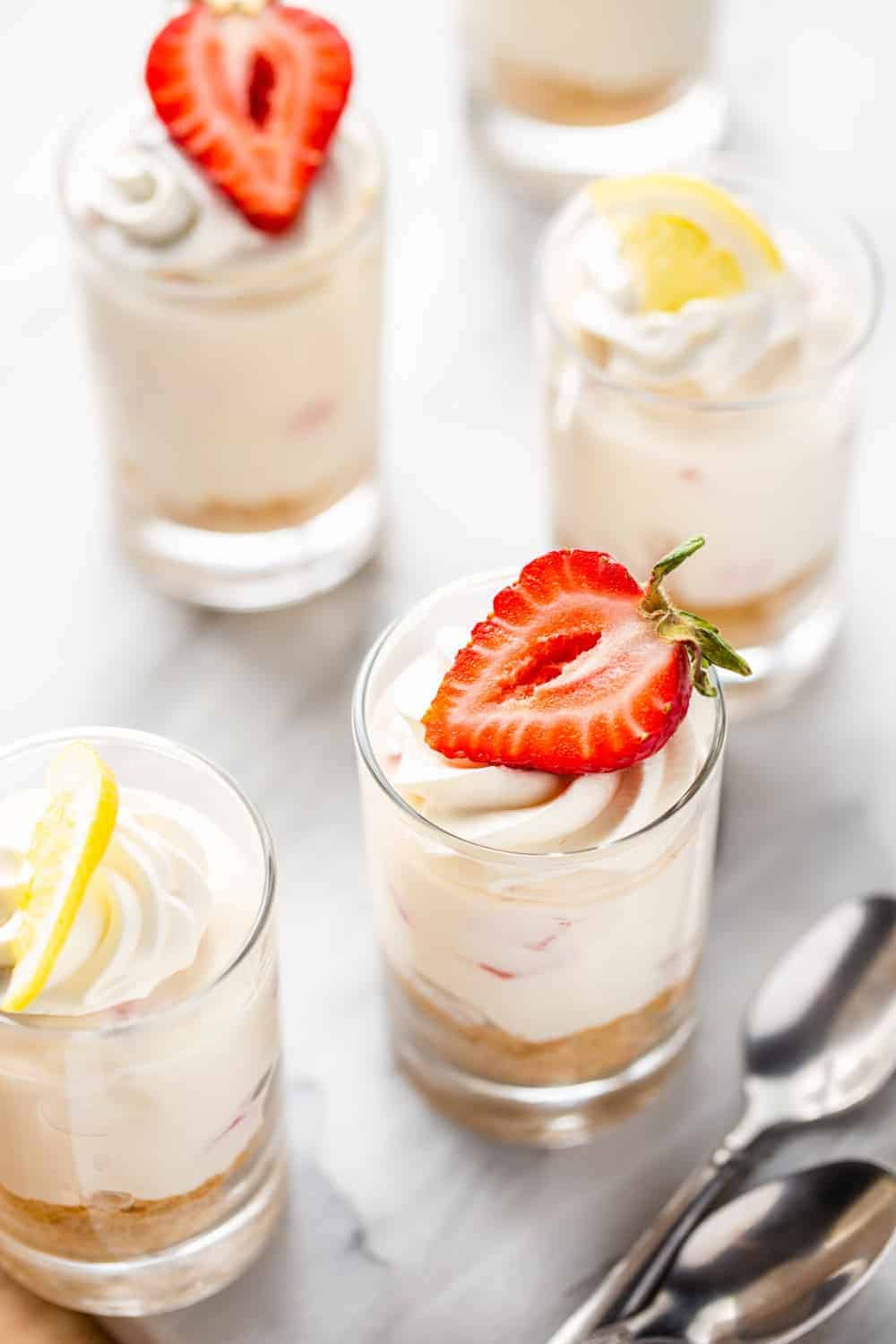 Mini no-bake strawberry lemonade cheesecakes in shot glasses, topped with whipped cream and a mixture of lemons and strawberries