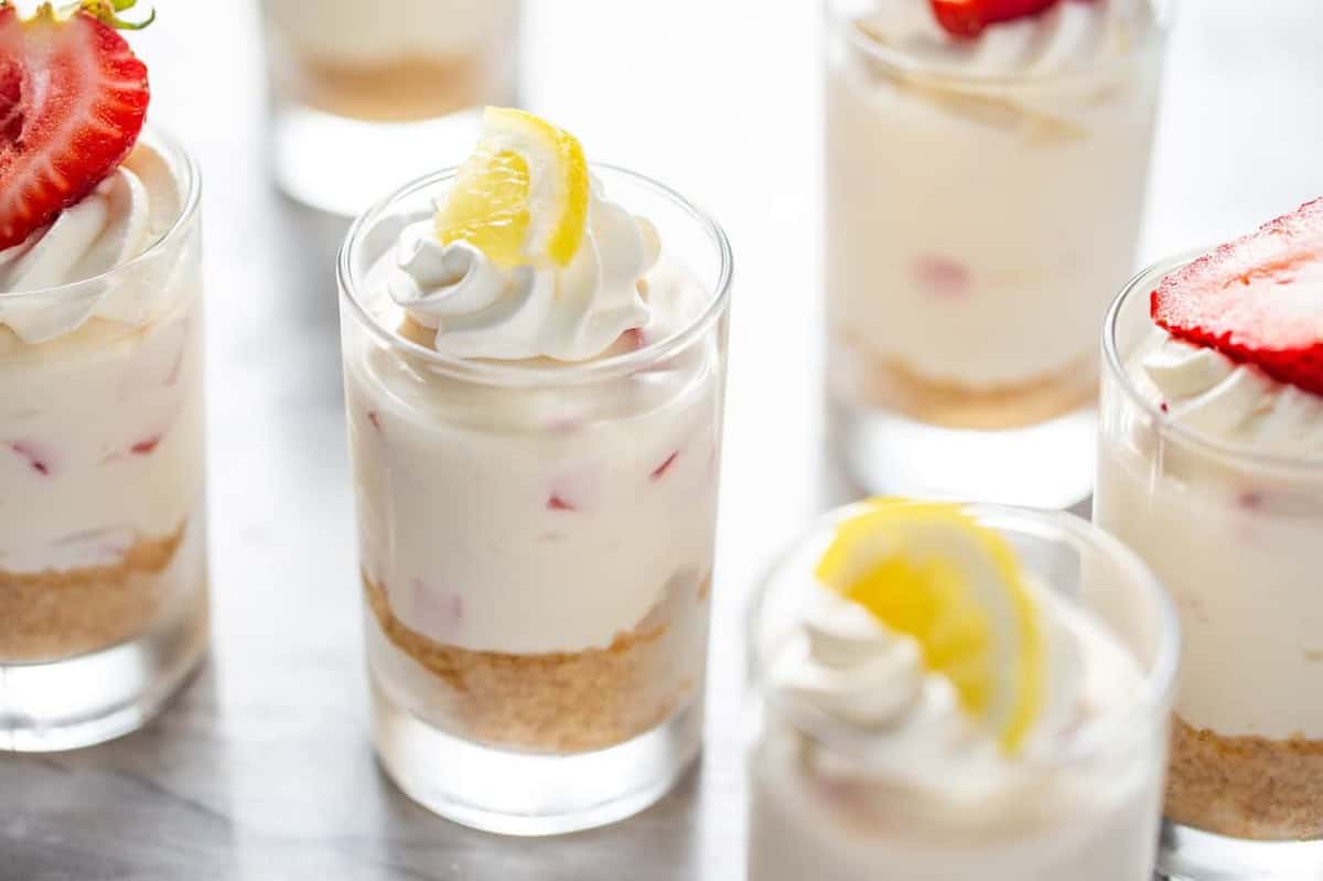 Assorted no-bake strawberry lemonade cheesecakes in dessert shot glasses, topped with whipped cream and fruit
