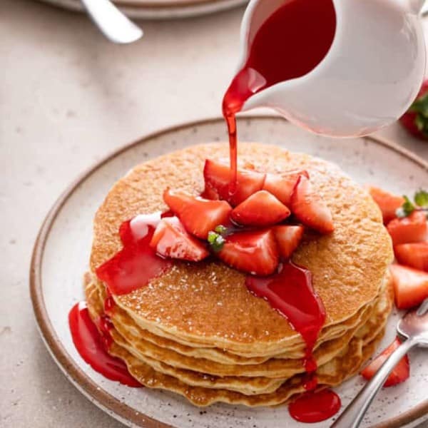 Stack of gluten-free pancakes topped with strawberries on a speckled plate. Strawberry syrup is being poured over the pancakes.
