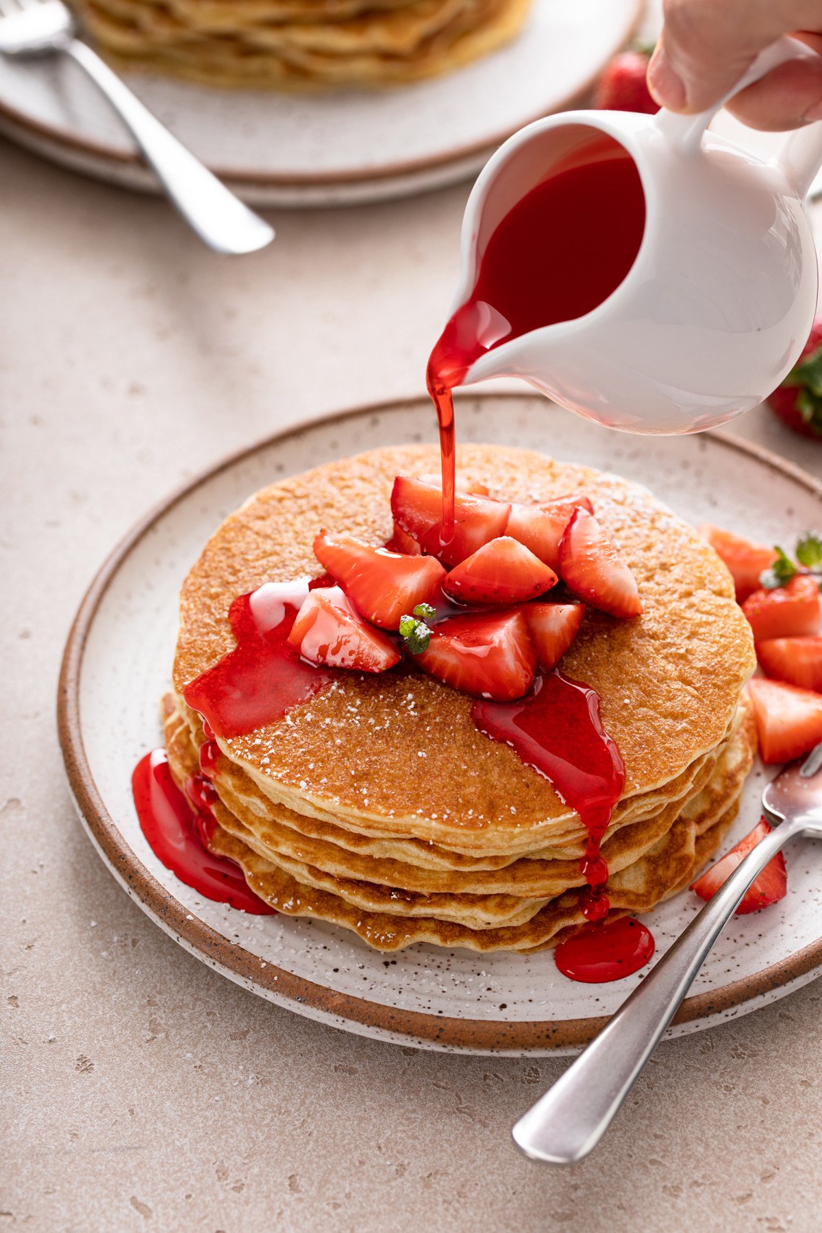 Stack of gluten-free pancakes topped with strawberries on a speckled plate. Strawberry syrup is being poured over the pancakes.
