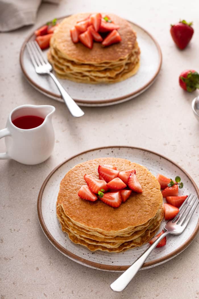 Two plates each holding a stack of gluten-free pancakes topped with fresh strawberries. A pitcher of strawberry syrup is nearby.