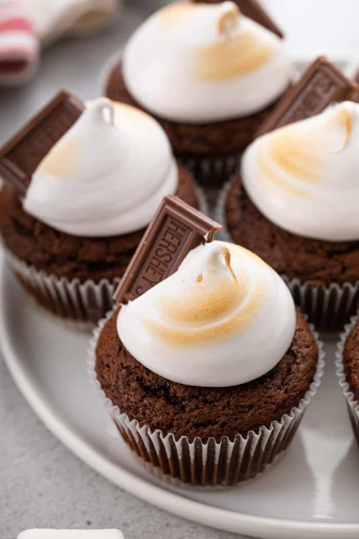 Close up of s'mores cupcakes topped with marshmallow frosting and a chocolate square on a white platter.