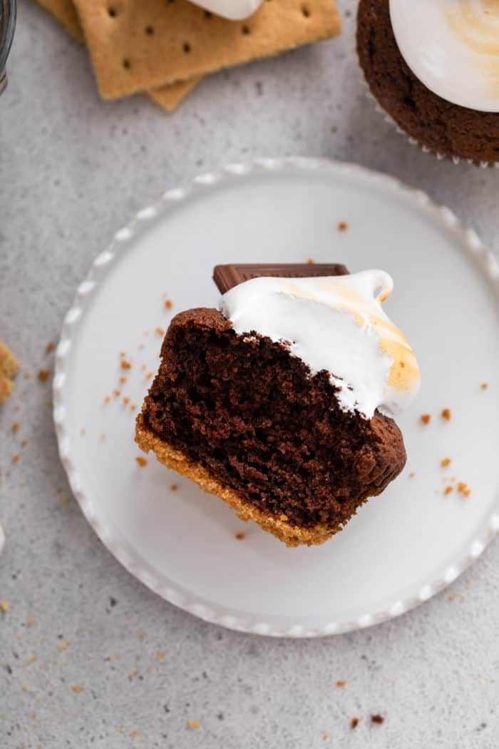 Halved s'mores cupcake laid on a white plate to show the graham cracker base, chocolate cake, and marshmallow frosting topping.