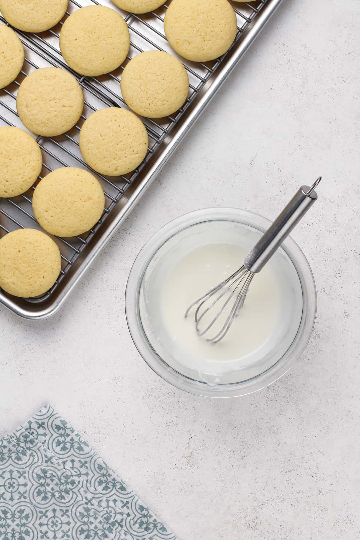 Lemon glaze in a glass bowl next to a cooling rack of cookies.