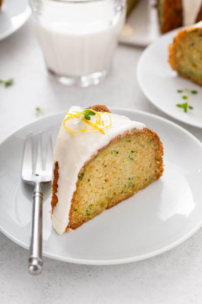 Slice of glazed lemon zucchini cake next to a fork on a white plate. A glass of milk is visible in the background.