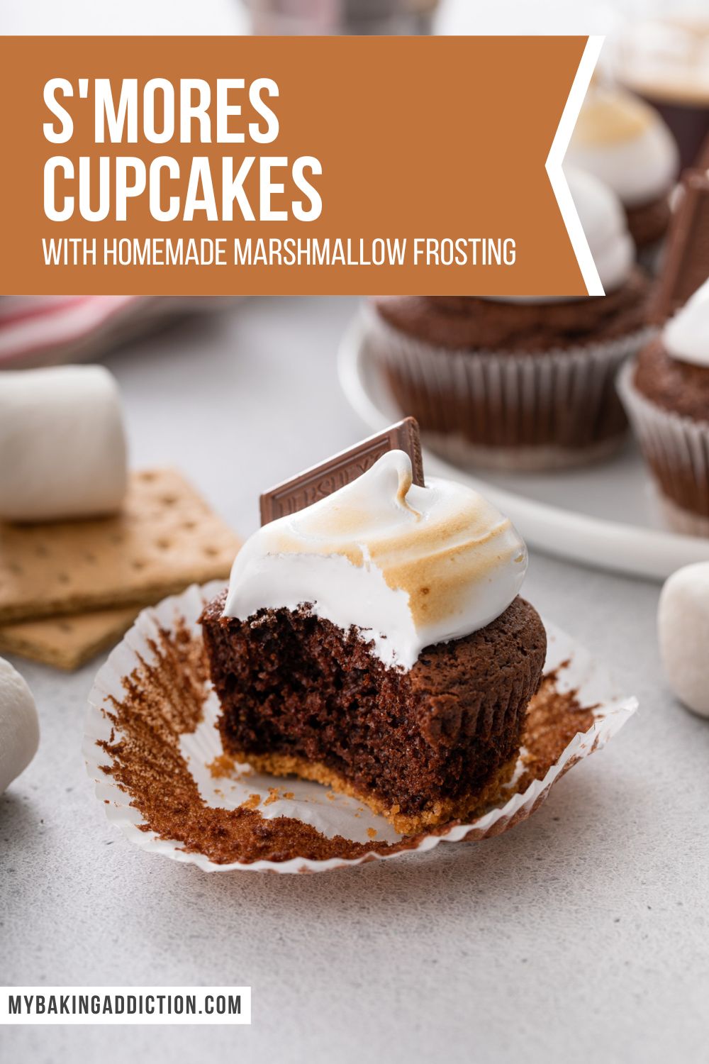 Unwrapped s'mores cupcake with a bite taken out of it. A platter of more cupcakes is visible in the background. Text overlay includes recipe name.