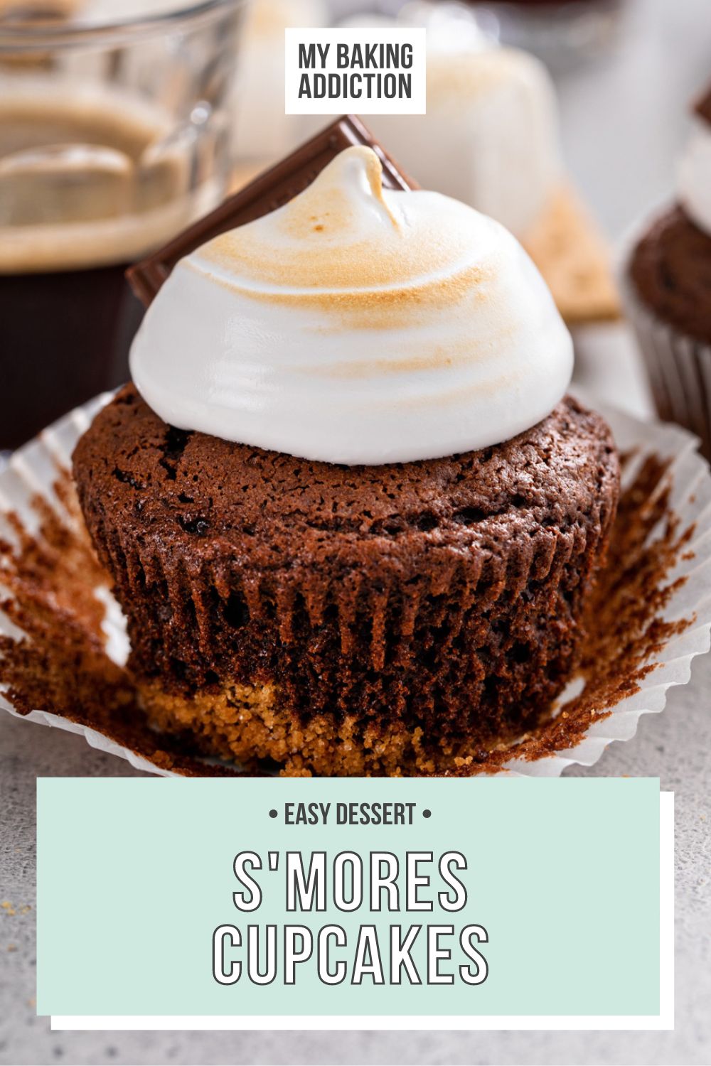 Unwrapped s'mores cupcake set on a gray countertop. A cup of espresso and more cupcakes are visible in the background. Text overlay includes recipe name.