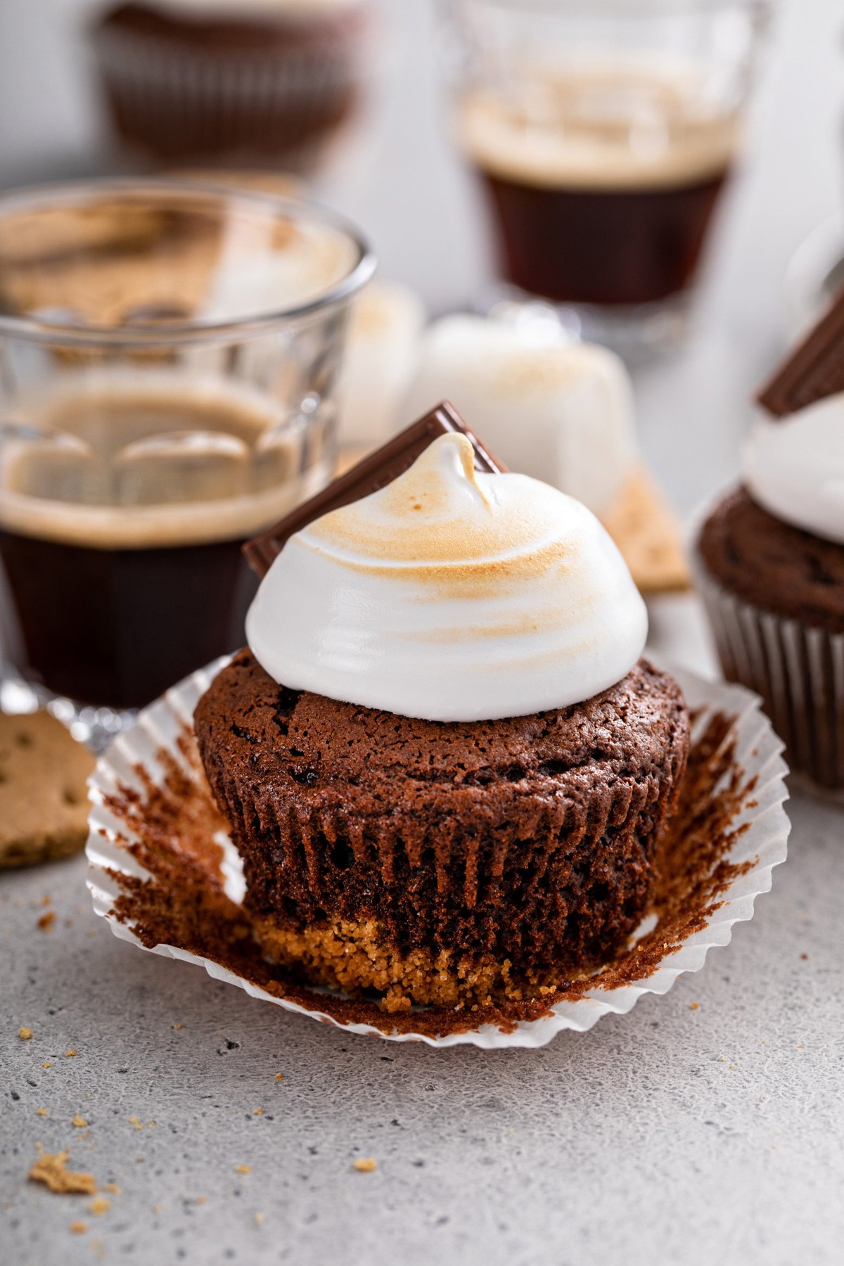 Unwrapped s'mores cupcake set on a gray countertop. A cup of espresso and more cupcakes are visible in the background.