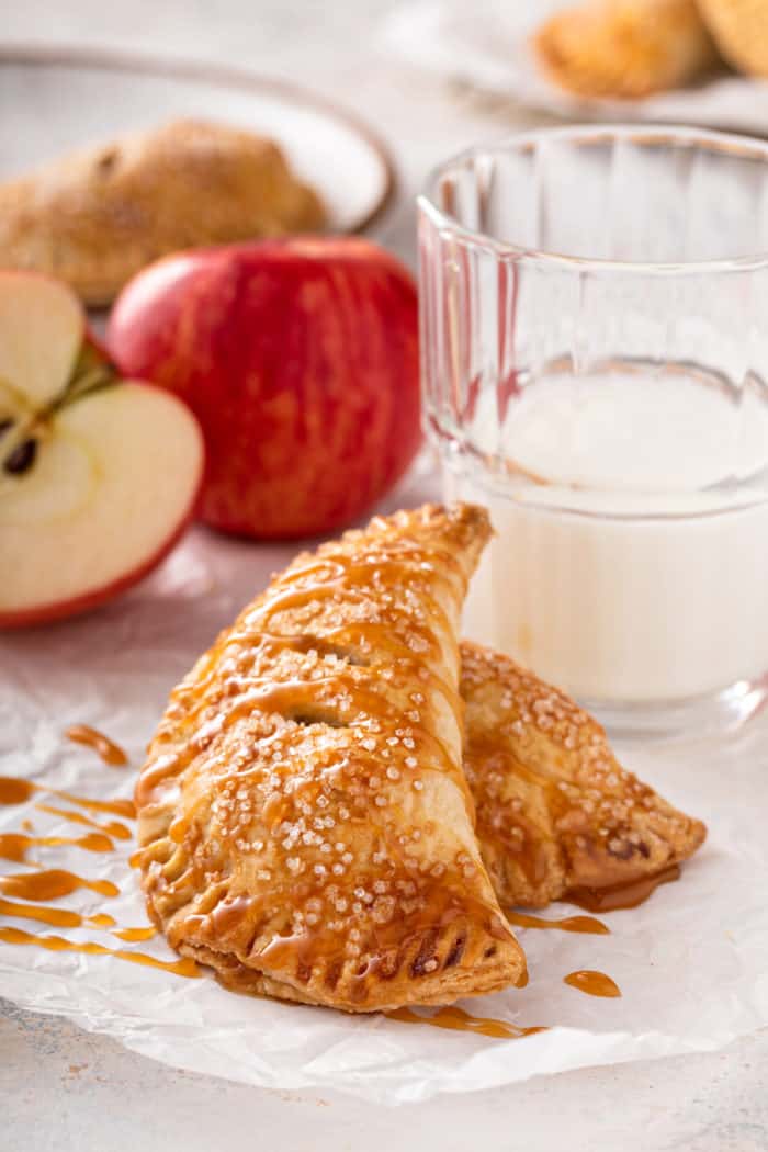 Two apple hand pies drizzled in caramel sauce, set in front of a glass of milk.