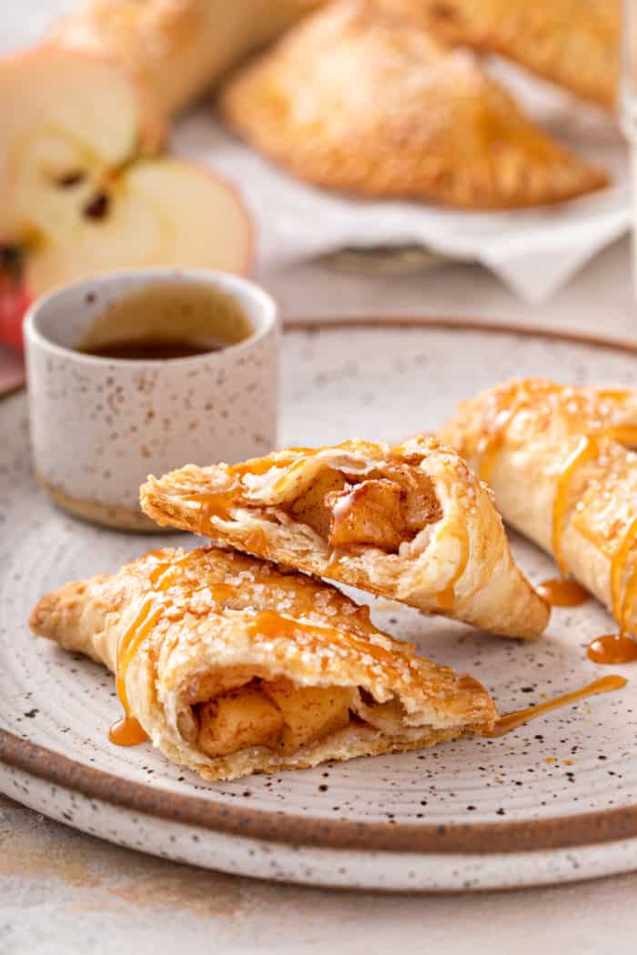 Two apple hand pies on a plate. One of them is cut in half and drizzled with caramel sauce.