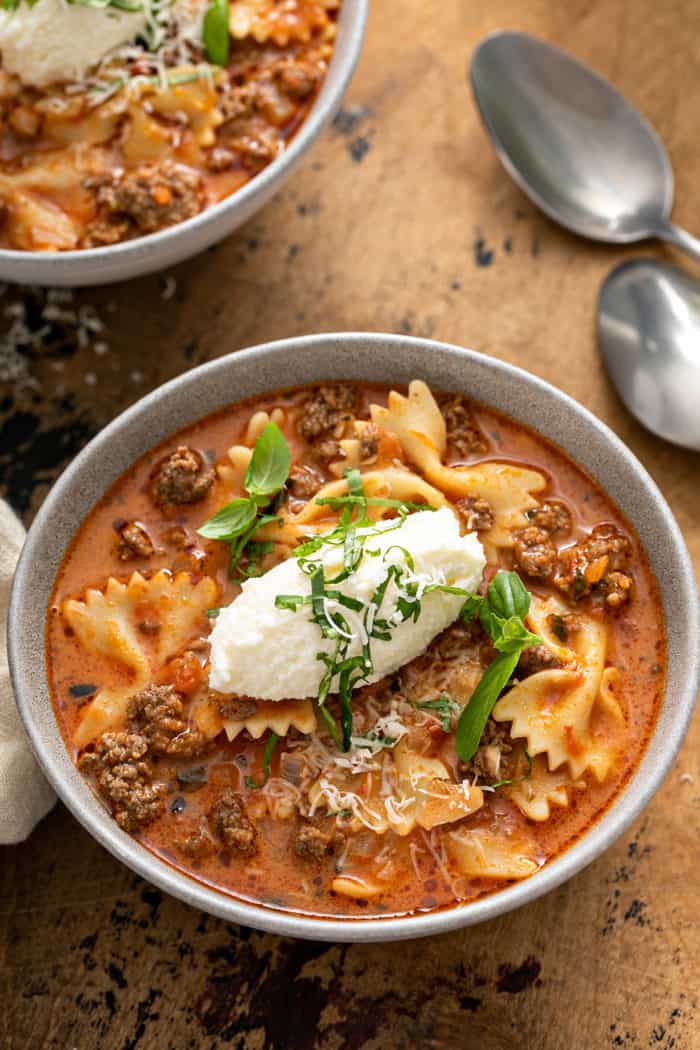 Close up of lasagna soup in a white bowl, topped with a spoonful of ricotta cheese