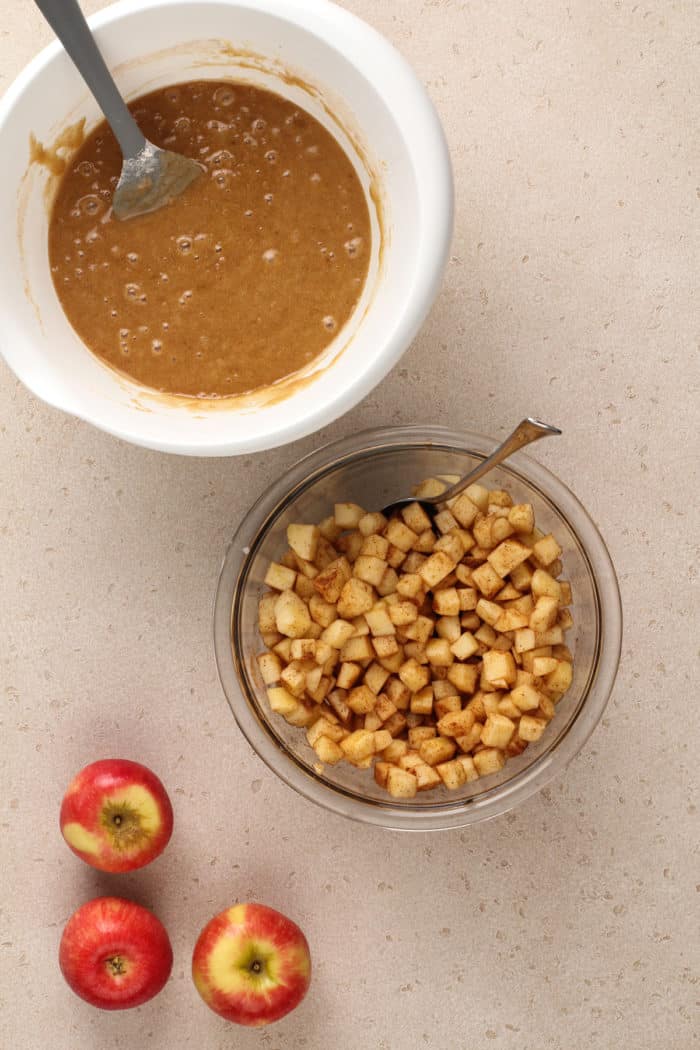 Cake batter in a white bowl, set next to cinnamon-coated apples in a glass bowl.