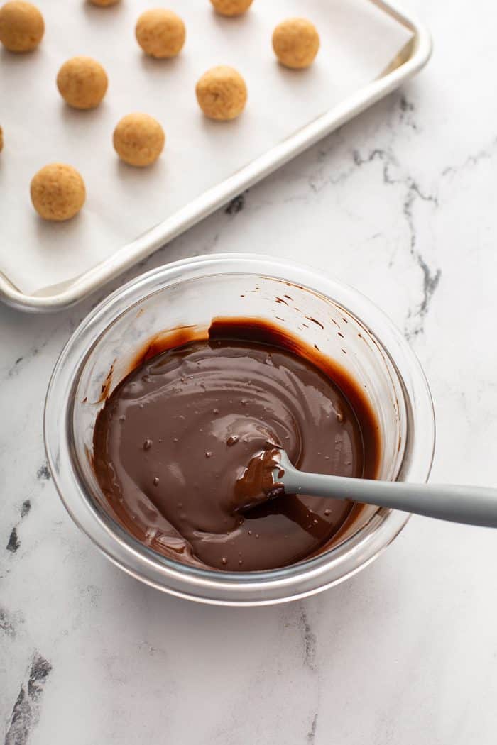 spatula stirring melted chocolate in a small glass bowl next to a tray of shaped cookie balls
