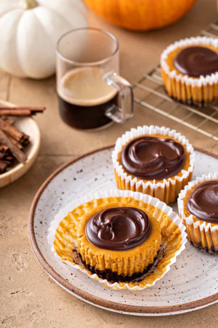 Three mini pumpkin cheesecakes on a speckled plate. The front cheesecake is unwrapped. All of them are topped with chocolate ganache.