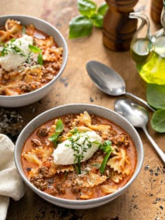 Two bowls of lasagna soup on a wooden table, each topped with a dollop of ricotta cheese and basil