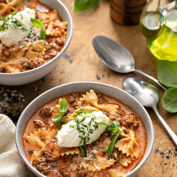 Two bowls of lasagna soup on a wooden table, each topped with a dollop of ricotta cheese and basil