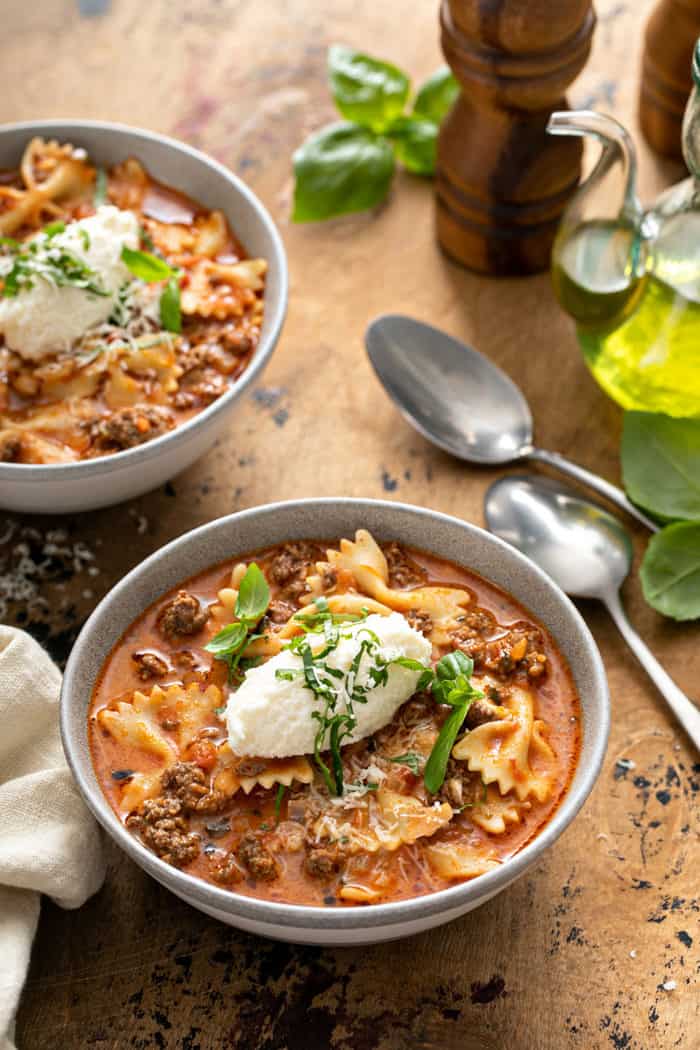 Two bowls of lasagna soup on a wooden table, each topped with a dollop of ricotta cheese and basil