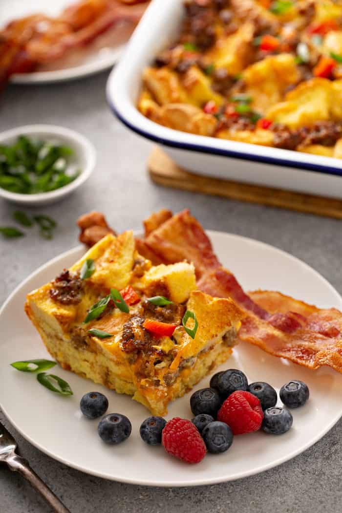 Slice of make-ahead breakfast casserole next to a slice of bacon and fresh berries on a white plate. A casserole dish of more breakfast casserole is in the background