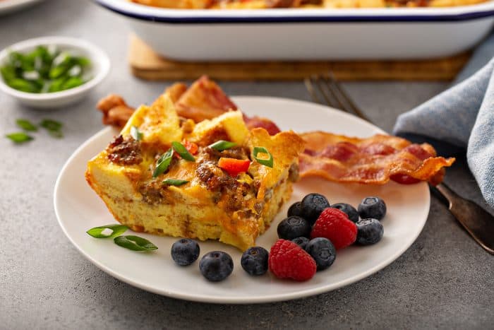 Side view of a slice of breakfast casserole next to bacon and fresh berries on a white plate with a casserole dish in the background