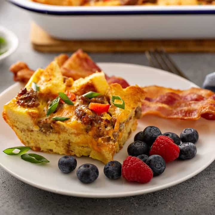 Side view of a slice of breakfast casserole next to bacon and fresh berries on a white plate with a casserole dish in the background