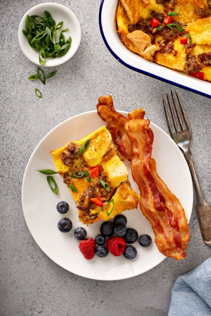 Overhead view of a slice of make-ahead breakfast casserole, a piece of bacon, and fresh berries on a white plate set next to a fork