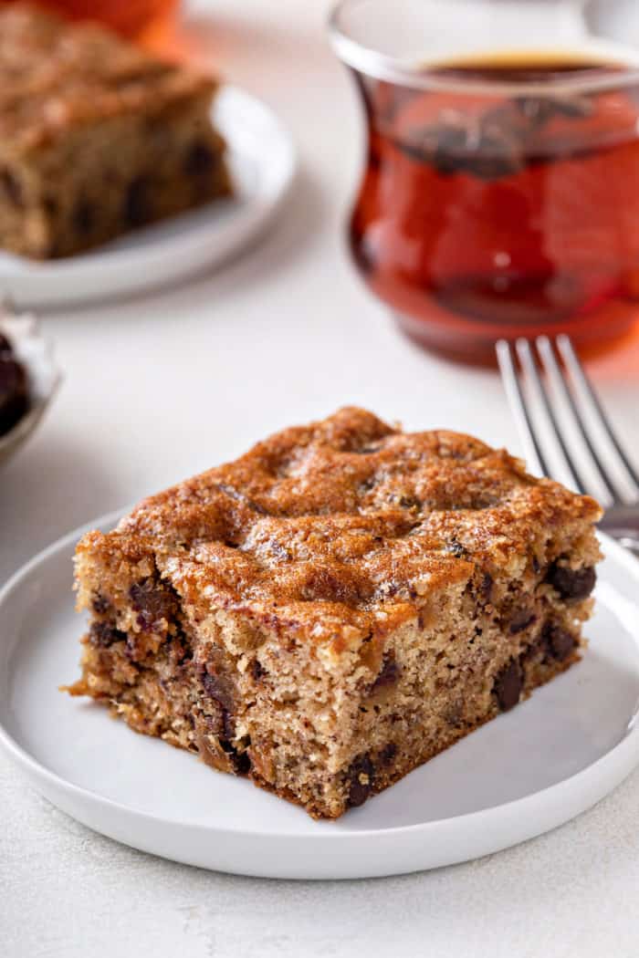 Large slice of date cake on a white plate with a cup of tea in the background.