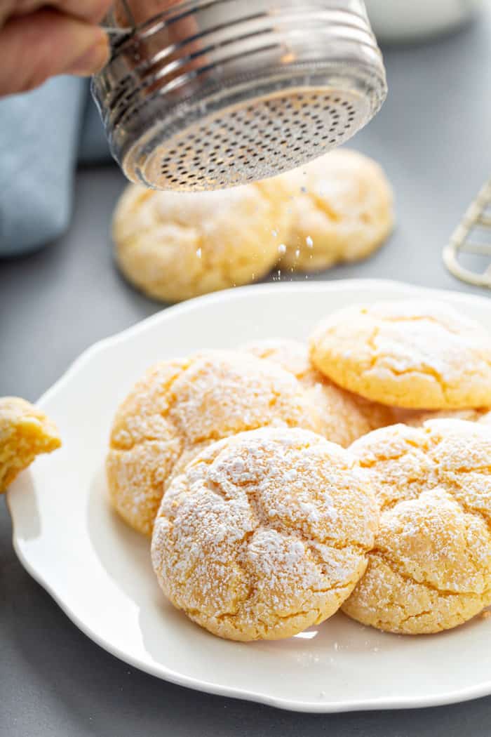 Powdered sugar being dusted on top of a pile of gooey butter cookies on a white plate