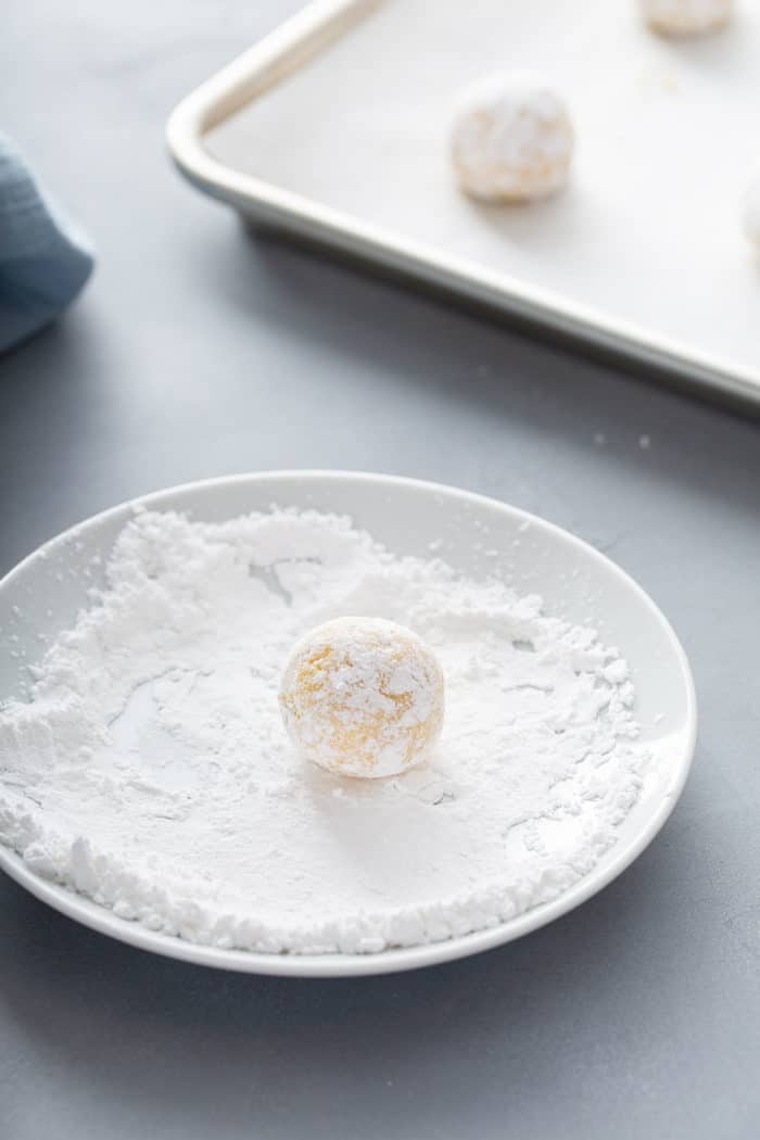 Ball of gooey butter cookie dough being rolled in powdered sugar on a white plate