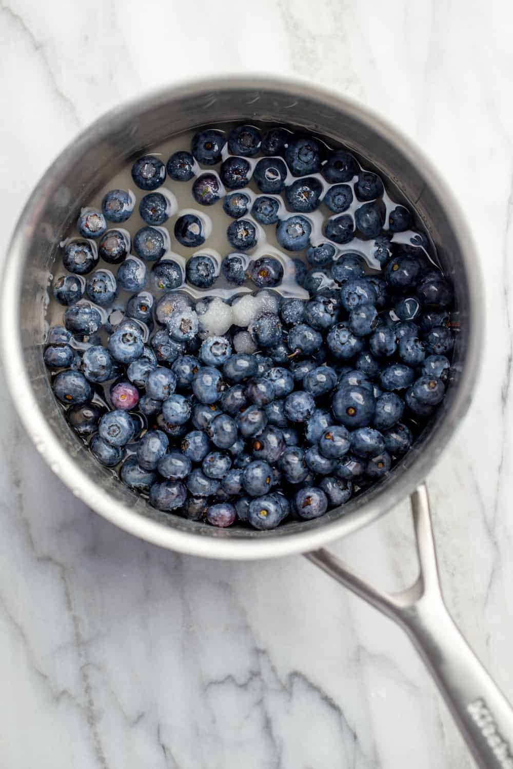Fresh blueberries, water and sugar in a saucepan on a marble surface