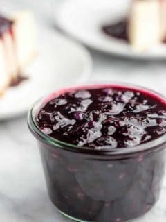 Homemade blueberry sauce in a glass jar with slices of cheesecake in the background