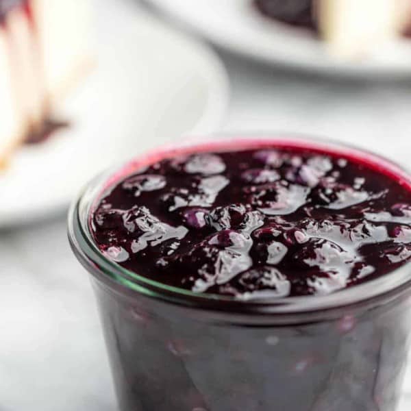 Homemade blueberry sauce in a glass jar with slices of cheesecake in the background