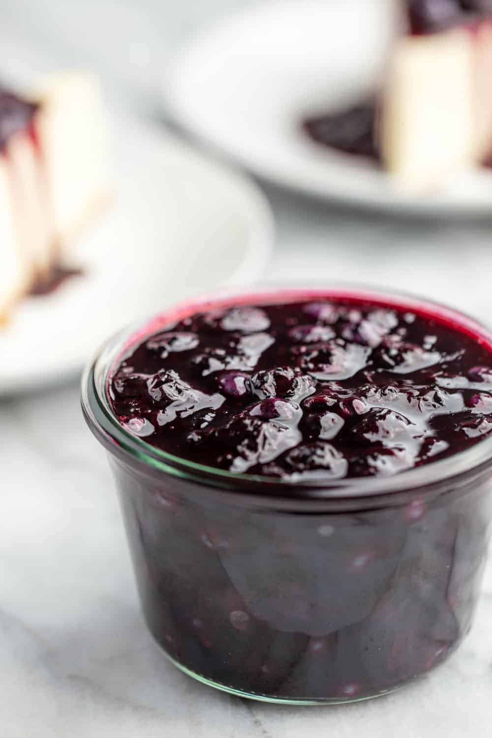 Homemade blueberry sauce in a glass jar with slices of cheesecake in the background