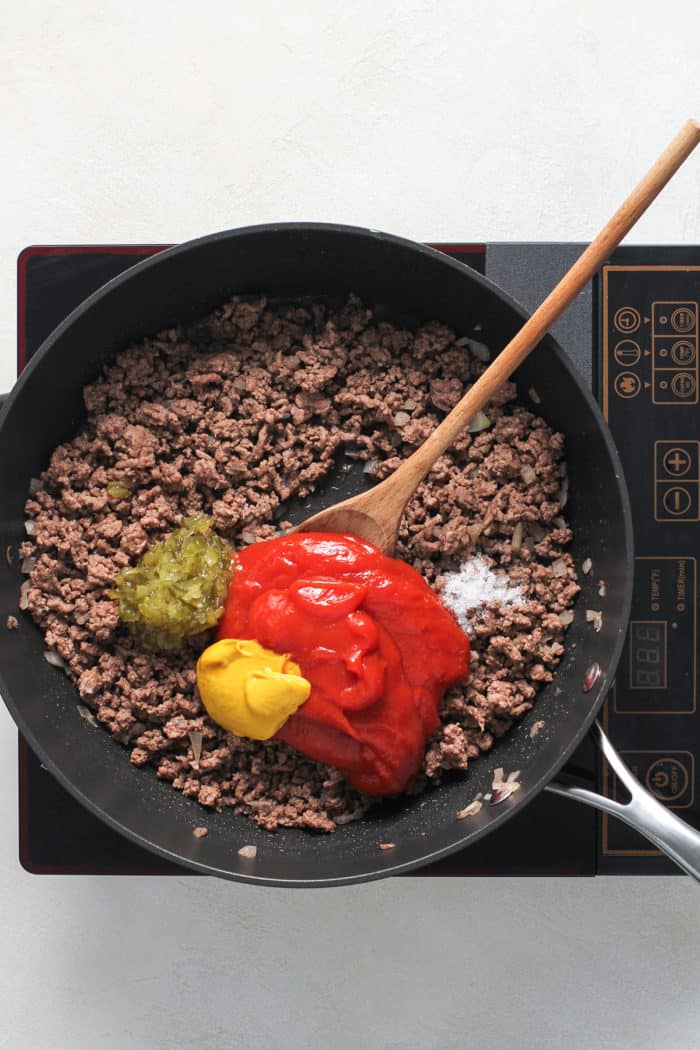 Ground beef in a skillet with ketchup, mustard, and relish being added