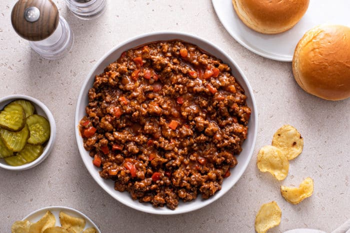 Sloppy joe mix in a white serving bowl, next to a plate of buns and a bowl of pickles.