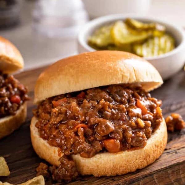 Close up of a sloppy joe sandwich on a wooden cutting board. A bowl of pickles is visible in the background.