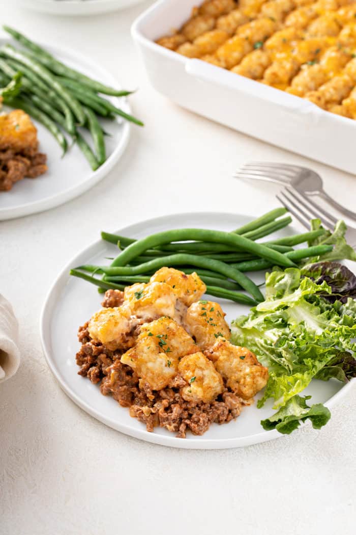 Serving of cheeseburger tater tot casserole on a white plate next to a salad and steamed green beans