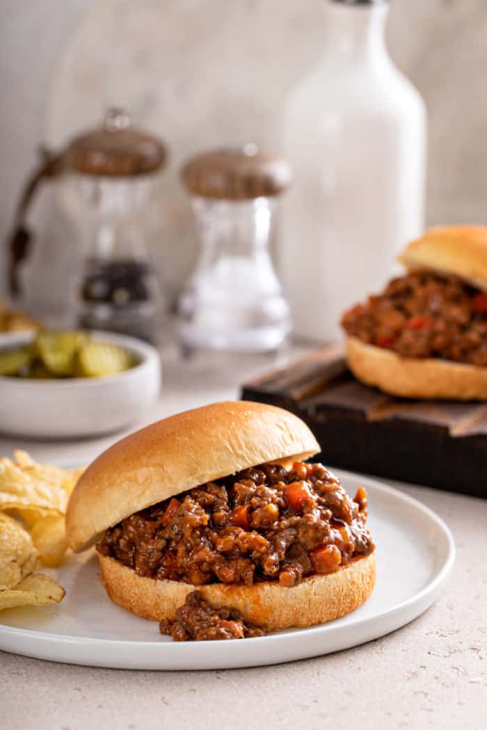 Sloppy joe sandwich on a hamburger bun, set on a white plate next to potato chips.