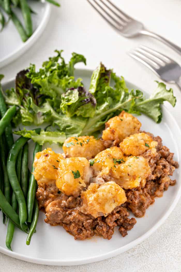 White plate with cheeseburger tater tot casserole next to green beans and salad