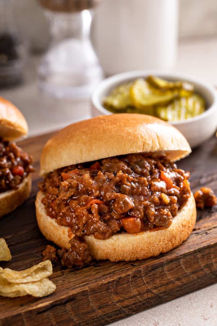 Close up of a sloppy joe sandwich on a wooden cutting board. A bowl of pickles is visible in the background.