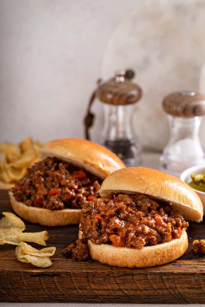 Two sloppy joes on hamburger buns, set on a wooden cutting board next to potato chips.