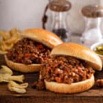 Two sloppy joe sandwiches on a wooden board next to potato chips.