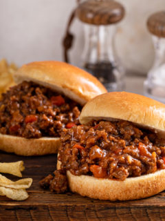 Two sloppy joe sandwiches on a wooden board next to potato chips.