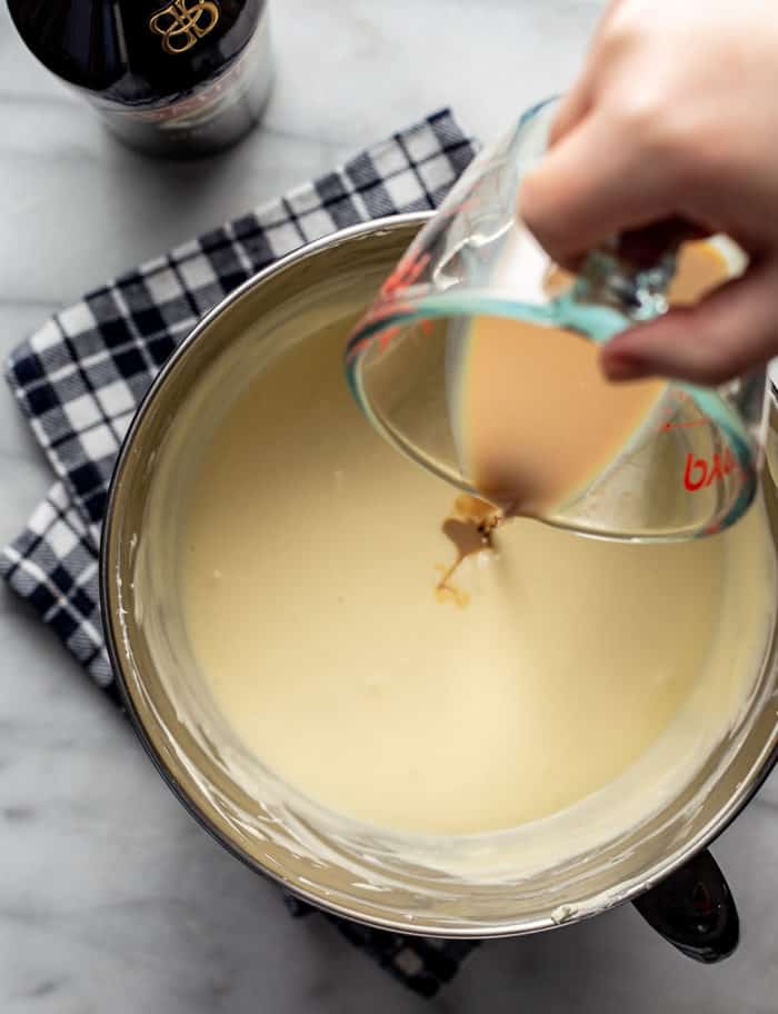 Baileys being poured into cheesecake filling batter in a metal mixing bowl
