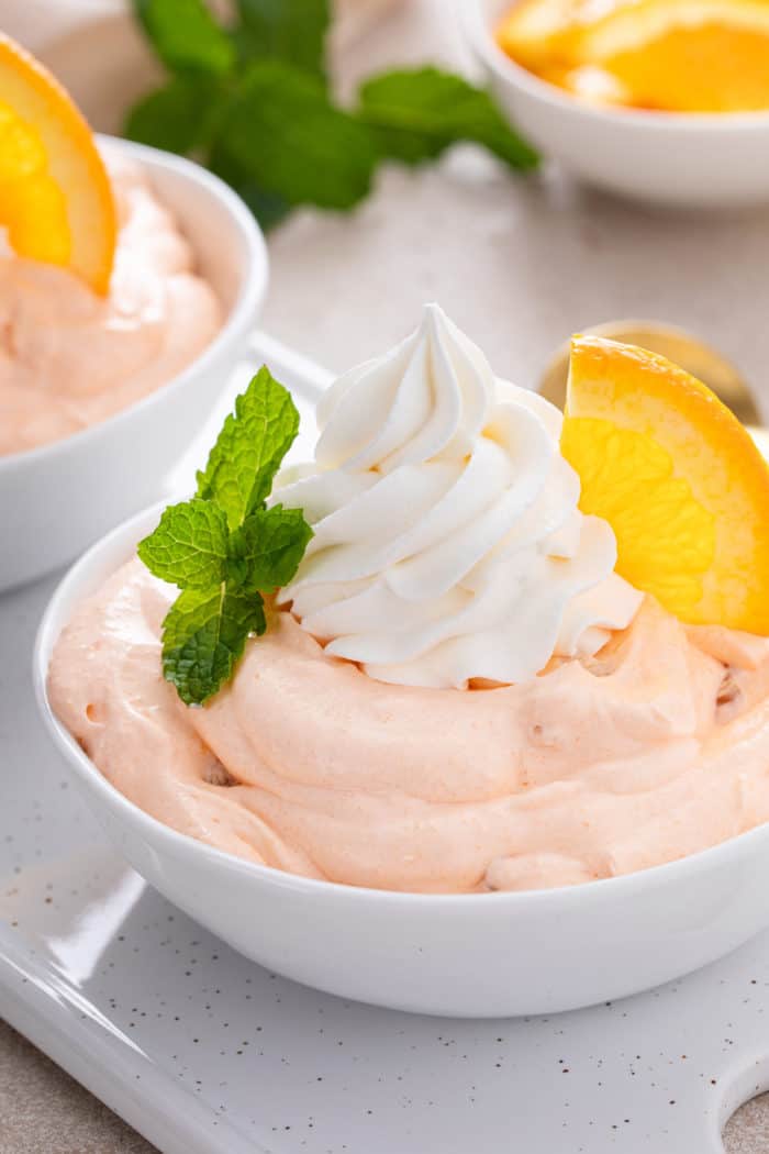 Close up of orange fluff in a white bowl, garnished with whipped cream and an orange slice.