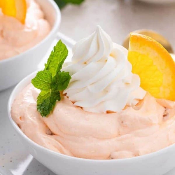 Close up of orange fluff in a white bowl, garnished with whipped cream and an orange slice.