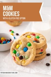 4 M&M cookies stacked on a piece of parchment paper in front of a glass of milk, with a 5th cookie leaning against the front of the stack. Text overlay includes recipe name.