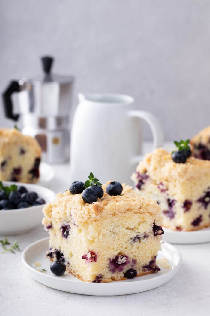 Plated slices of blueberry coffee cake on a countertop next to coffee creamer and a bowl of blueberries.