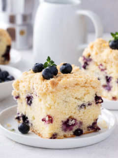 Slice of blueberry coffee cake on a white plate, with additional plated slices in the background.