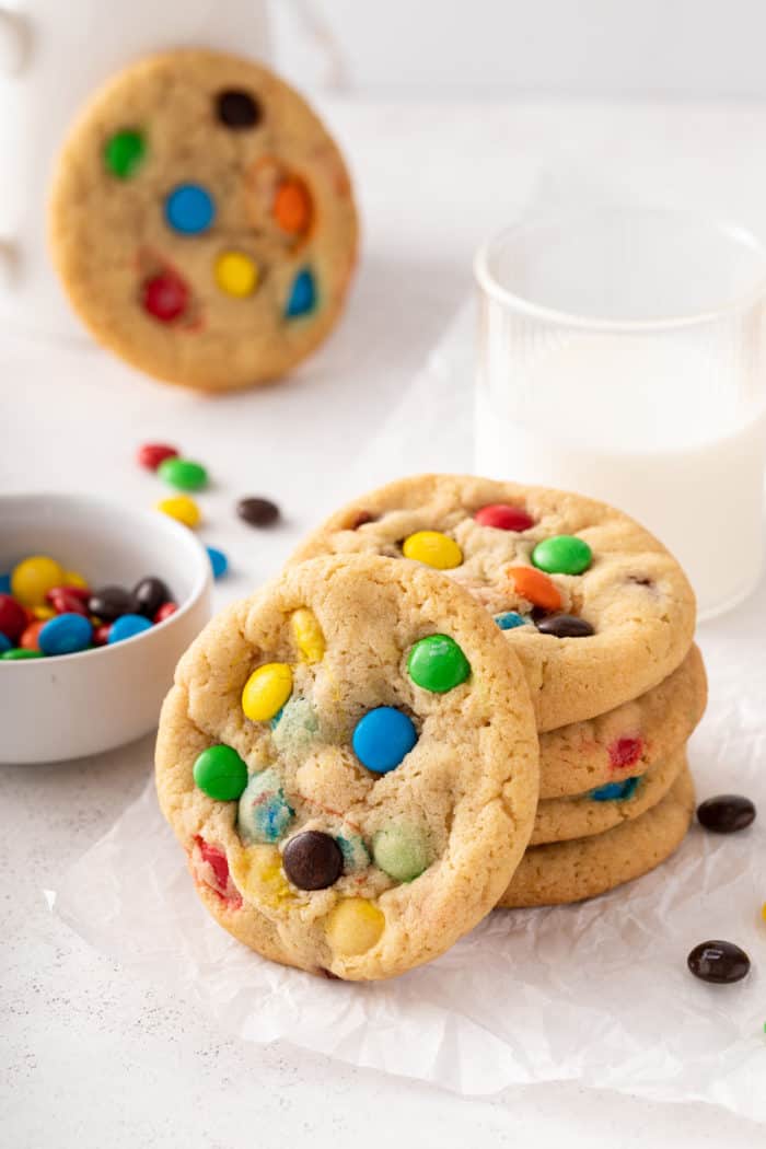 4 M&M cookies stacked on a piece of parchment paper in front of a glass of milk, with a 5th cookie leaning against the front of the stack.