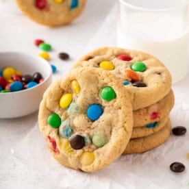 Stack of 4 m&m cookies on a piece of parchment paper with another cookie leaning against the stack.